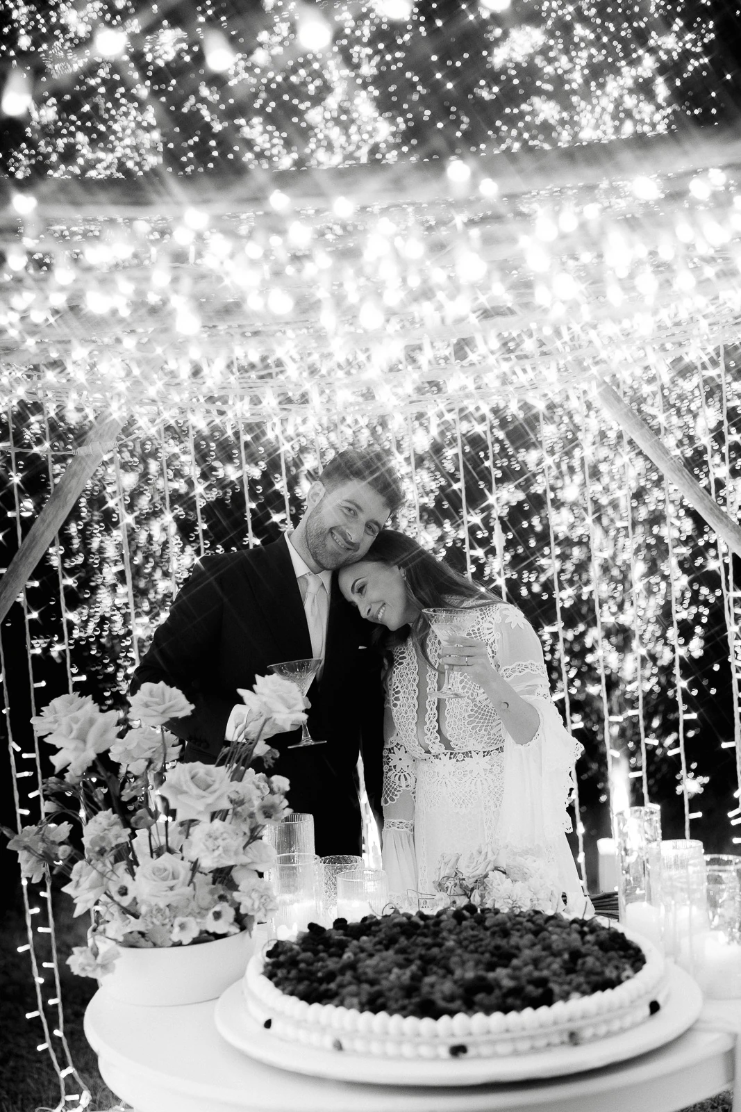 Beneath glowing string lights in Puglia, an elegant couple shares a quiet toast beside their wedding cake and seasonal blooms.