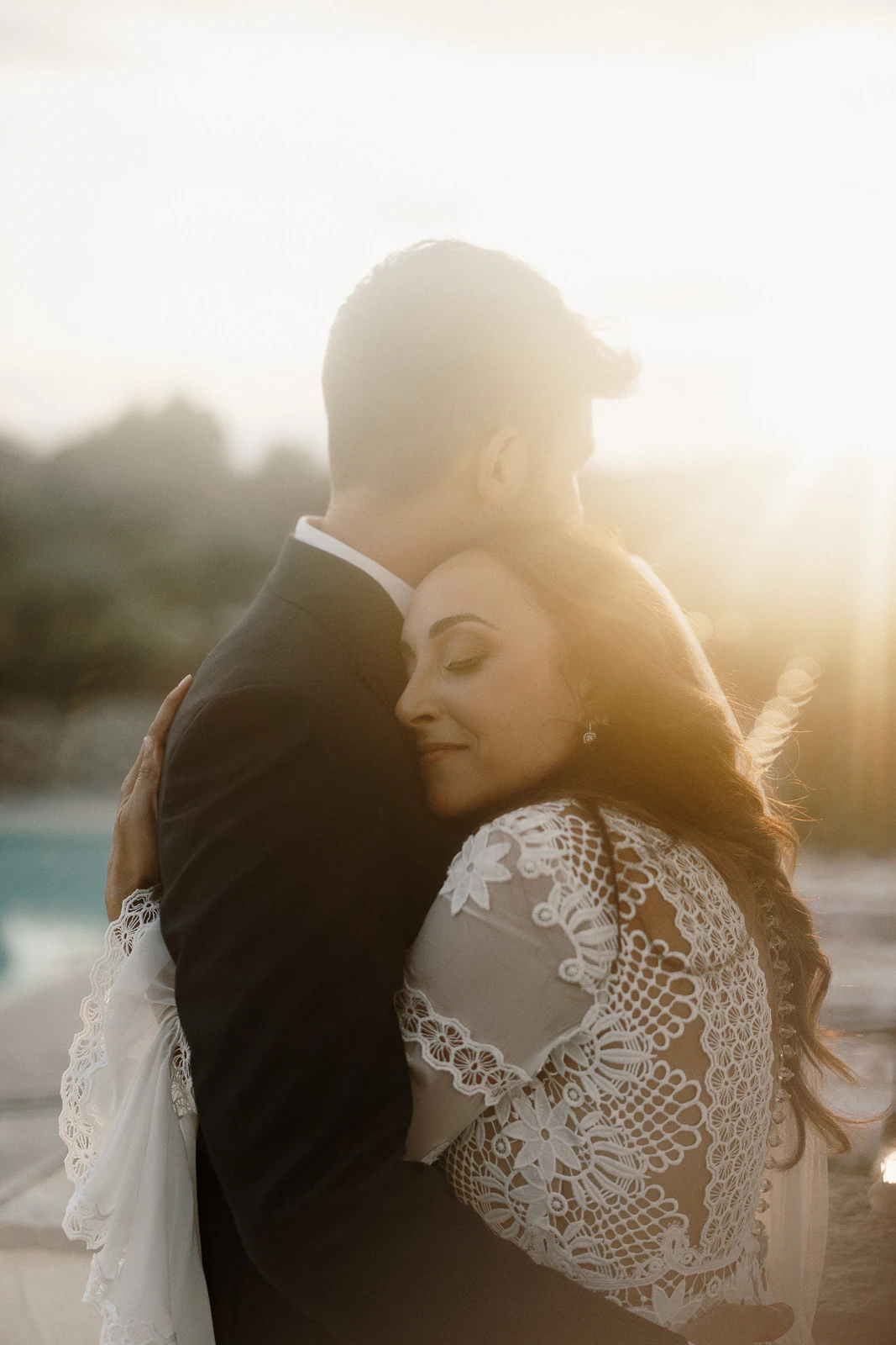 Embracing at sunset in Puglia, a woman in lace rests serenely against her partner, moments before golden Italian twilight.
