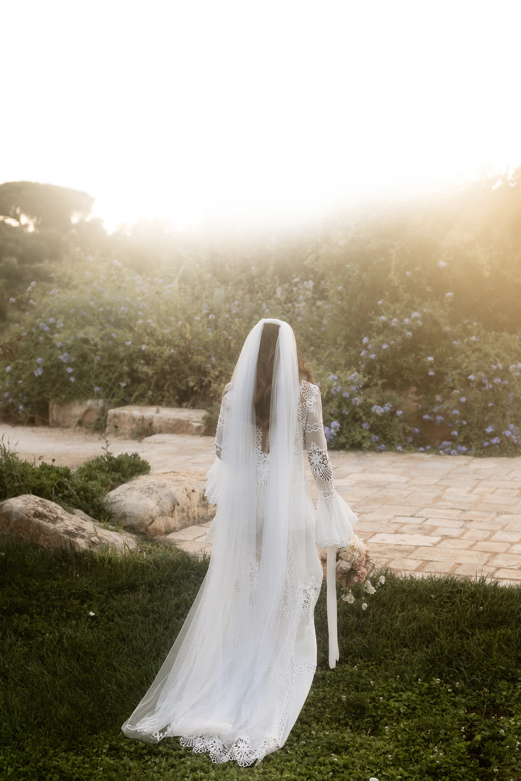 A bride in ethereal white lace gazes over a sun-dappled Italian garden, bouquet in hand, embodying Puglia romance.
