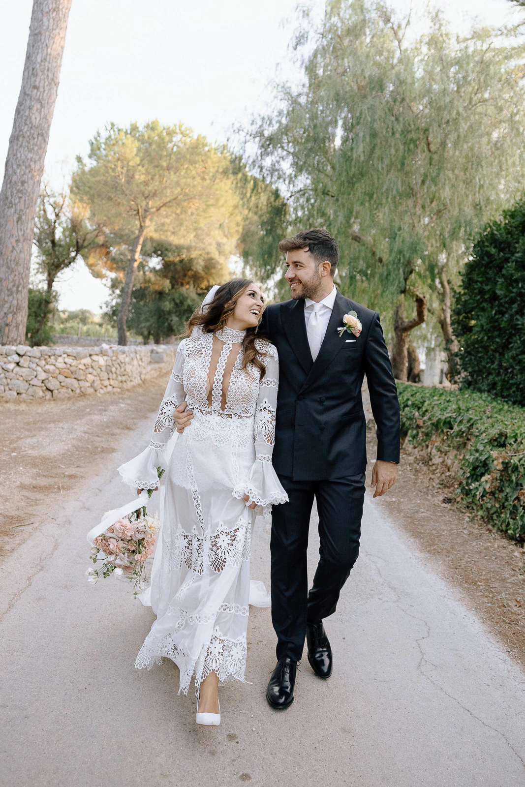 Amidst sunlit olive groves in Puglia, a joyful bride and groom stroll arm-in-arm, lost in each other’s gaze.