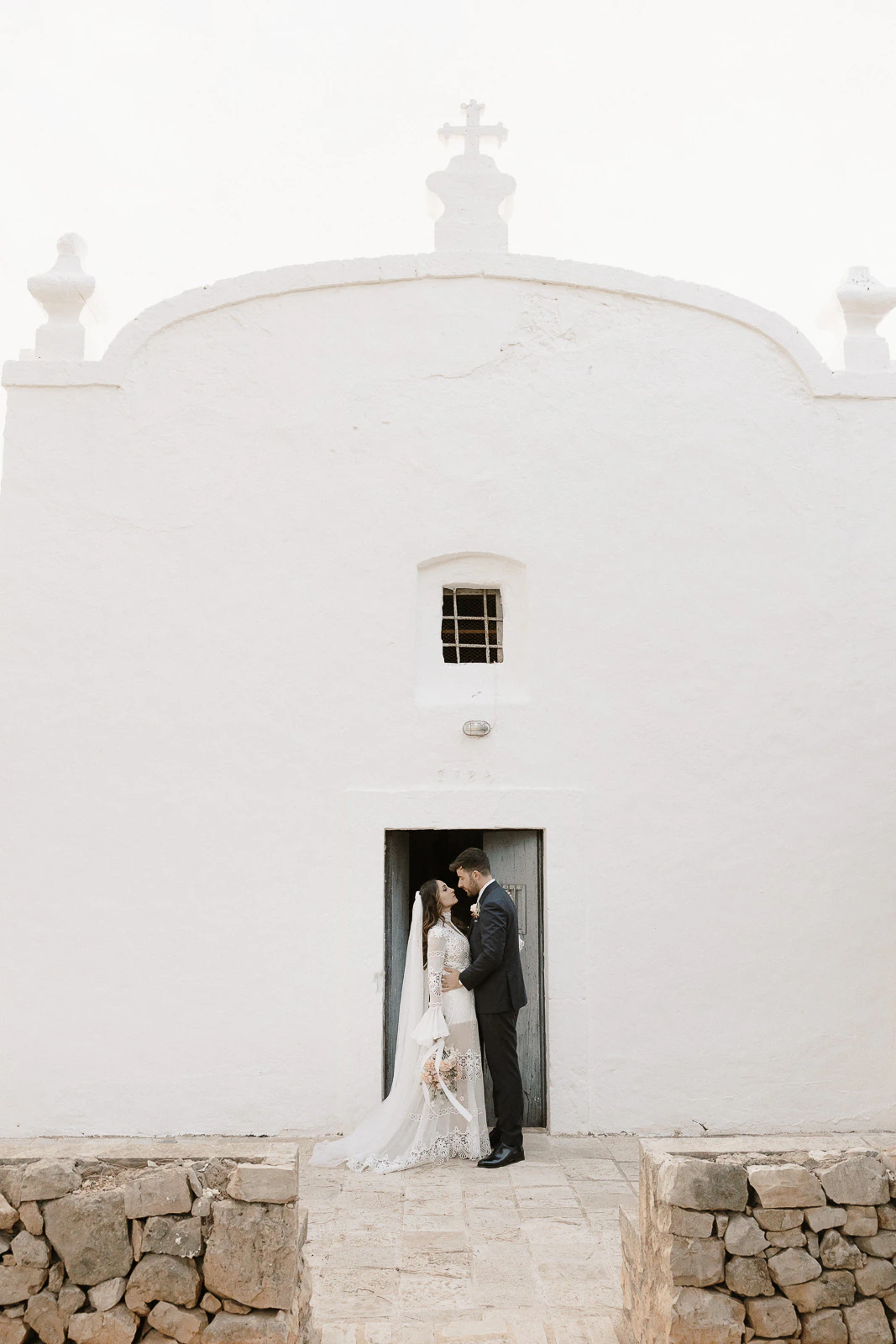 Bride and groom share a quiet moment in the doorway of a whitewashed Puglia chapel, sunlight illuminating their joyful expressions.