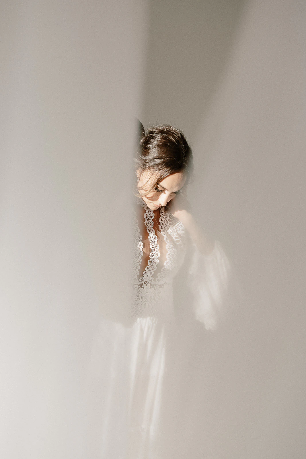 Bathed in ethereal light, a bride in delicate lace gazes through sheer curtains—romance lingers amid sunlit Puglia elegance.