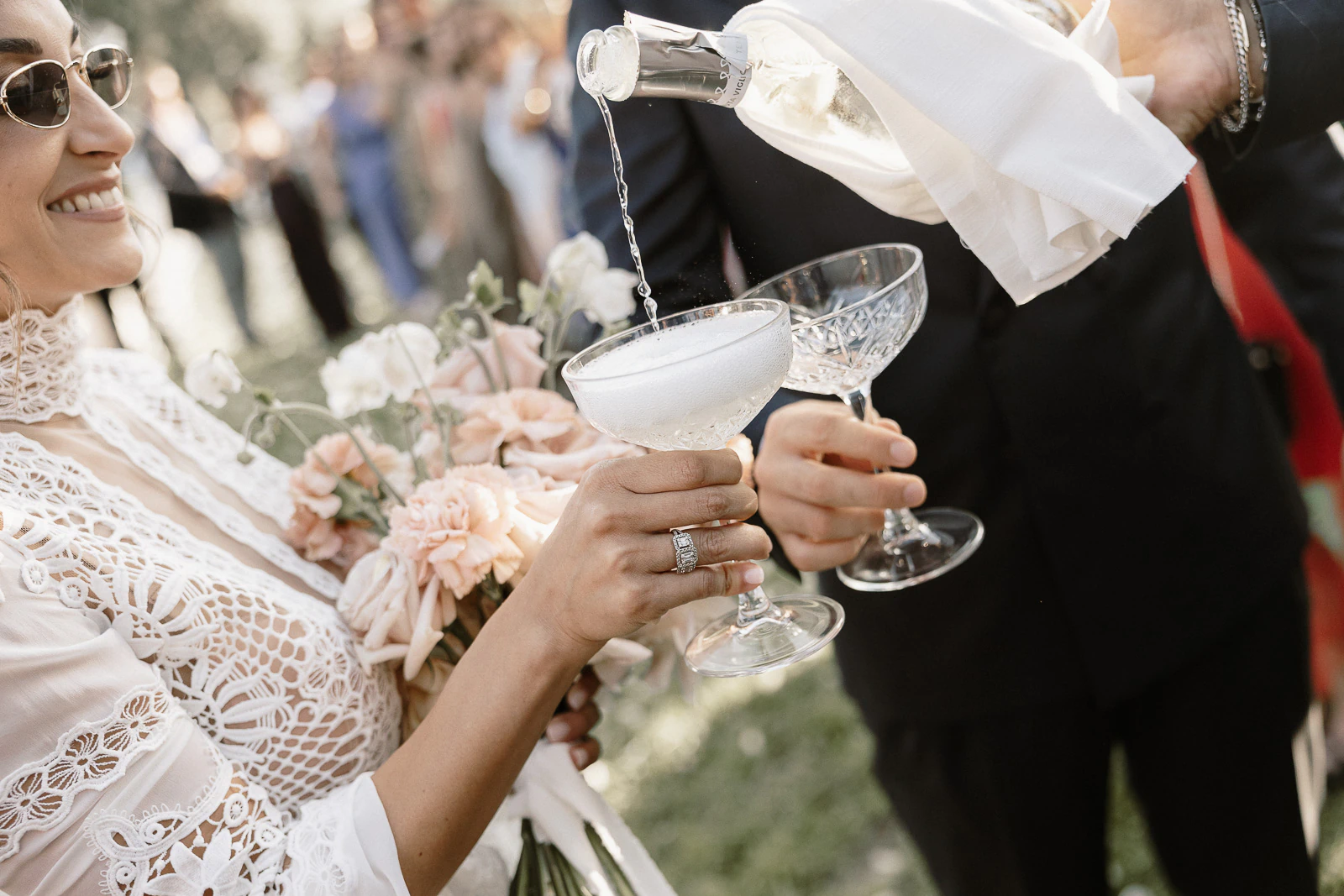 In sunlit Puglia, a radiant bride in lace smiles as sparkling wine is poured into coupe glasses, bouquet in hand.