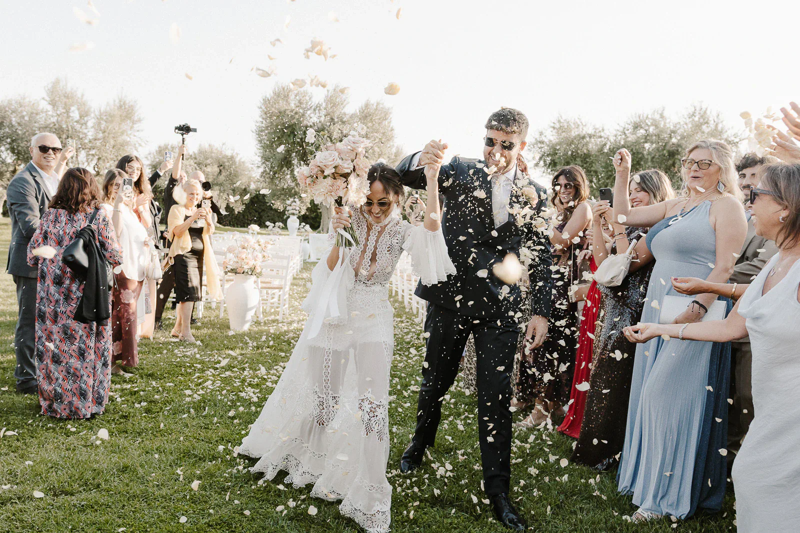 Radiant newlyweds stroll beneath a cascade of petals, guests rejoicing amid sunlit Puglia scenery and heartfelt celebration.