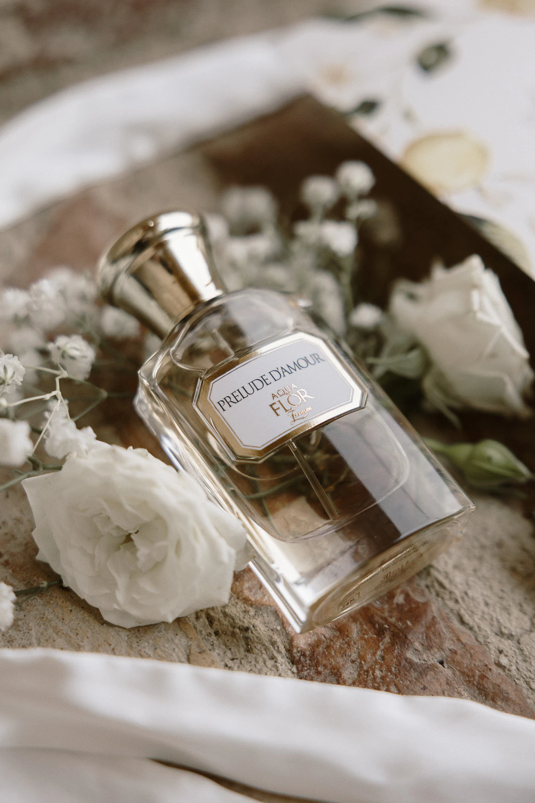PRELUDE D’AMOUR perfume rests amid delicate white blooms and lush greenery on a sunlit stone in romantic Puglia, Italy.