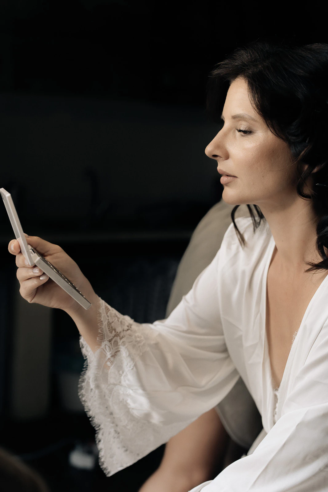 In Puglia, a bride-to-be in an elegant white robe softly smiles at her phone, anticipation filling the sunlit room.