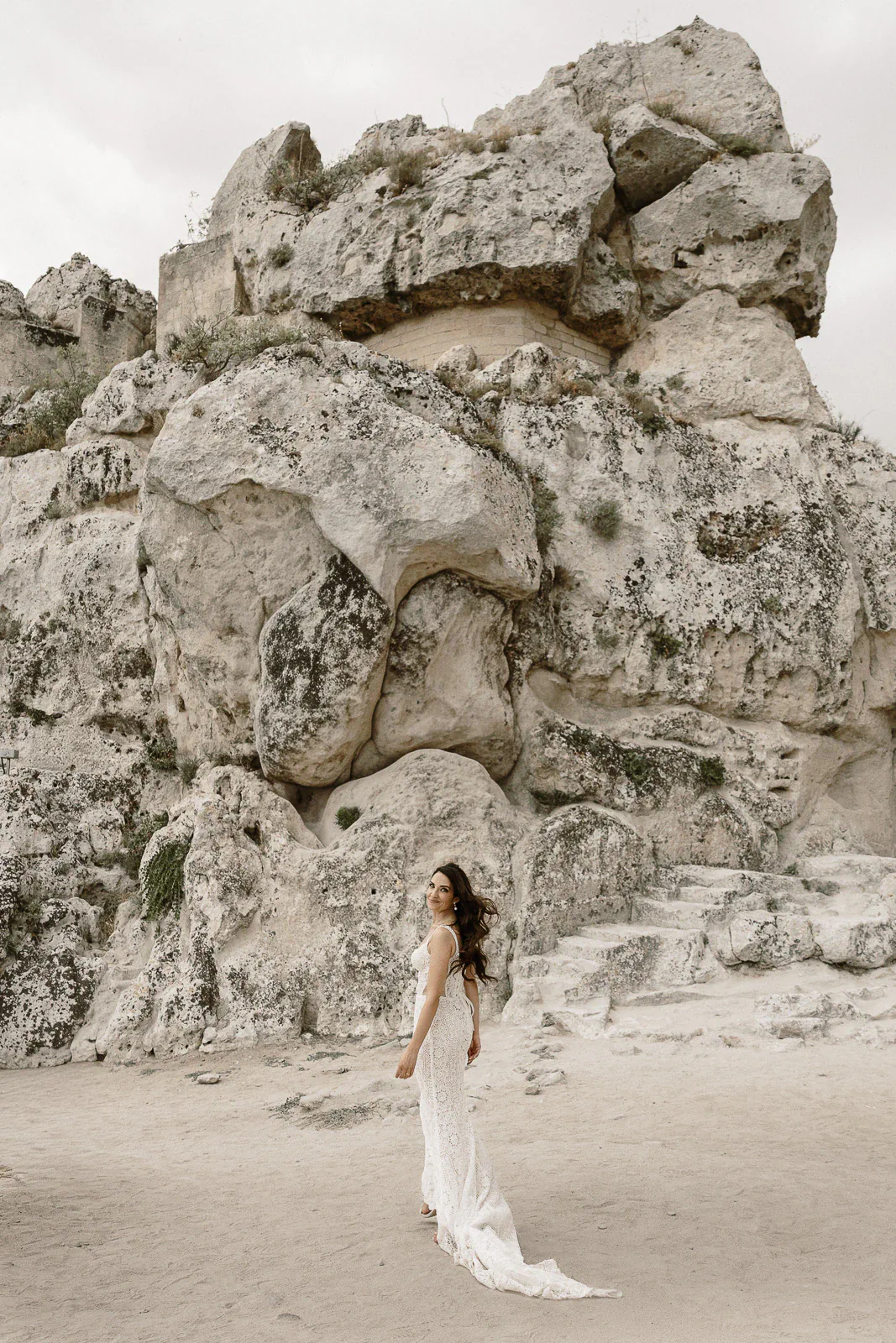 A bride in a delicate lace gown stands gracefully on sunlit sand amid dramatic rock formations, evoking romance in Puglia, Italy.