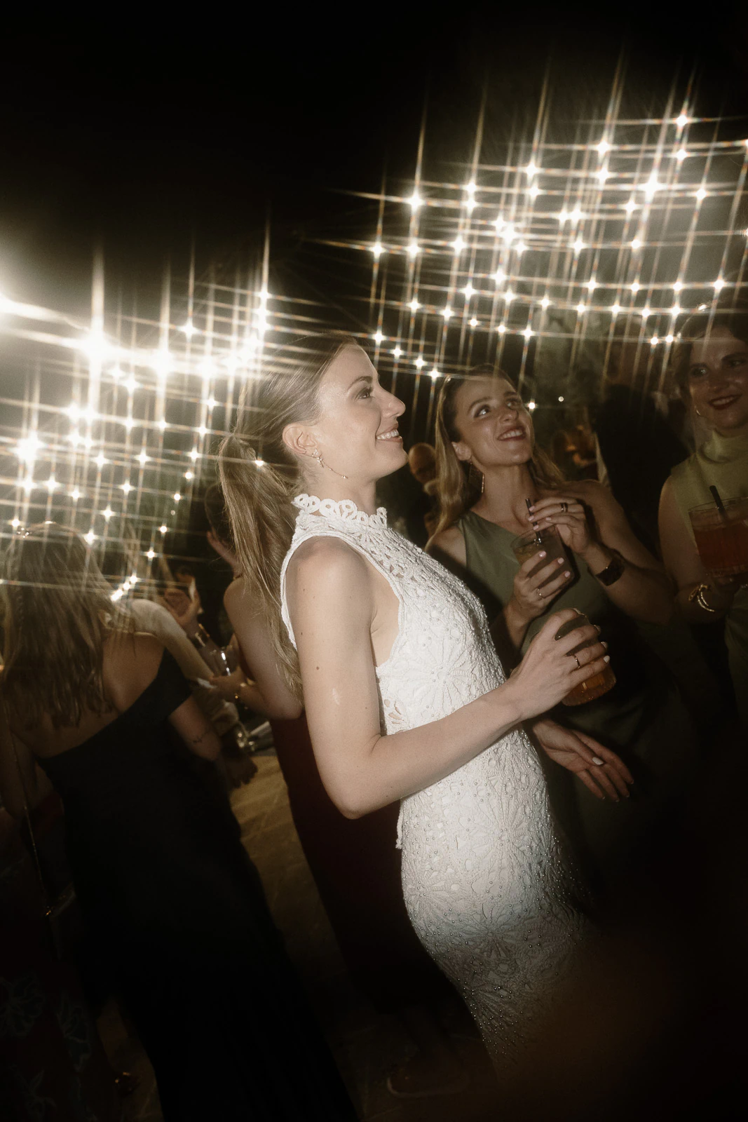 Elegant guests in evening gowns share a joyful toast beneath glowing string lights at a romantic Puglia wedding celebration.